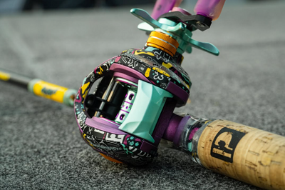 Colorful baitcasting fishing reel with graffiti-pattern housing and teal and purple accents, on cork handle labeled 'P'.