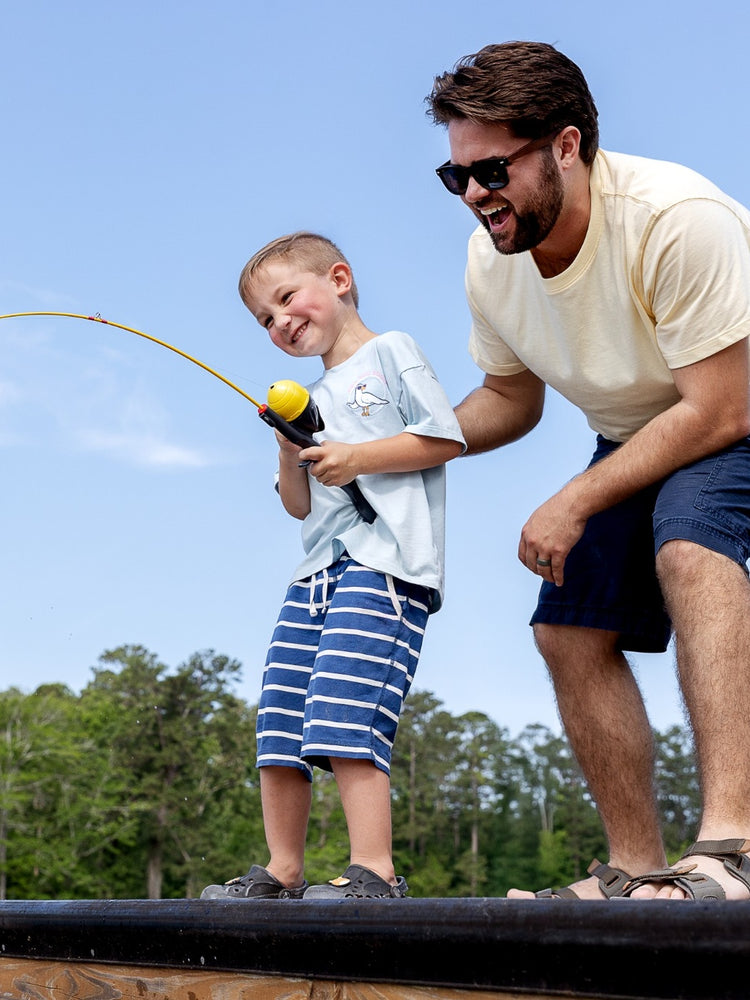 Father helping his young son reel in a fish under a clear blue sky.
