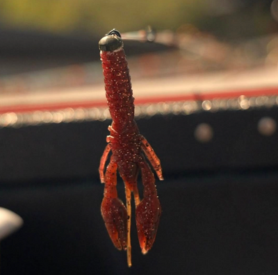 Red soft-plastic crawfish fishing lure hooked on a jig, hanging against a blurred boat background.