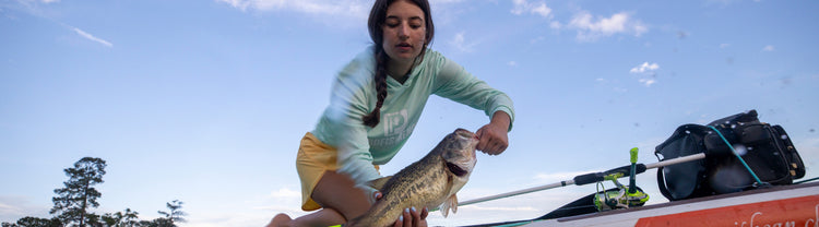 Young woman kneeling on a boat holding a large bass by the mouth and belly; fishing rod and gear visible.