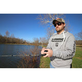 Man fishing by a pond wearing a gray hoodie labeled PROFISHIENCY and P, sunglasses and cap, holding a fishing rod.