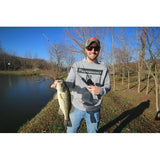 Man in PROFISHIENCY hoodie holding a largemouth bass by a pond, fishing rod in his other hand.